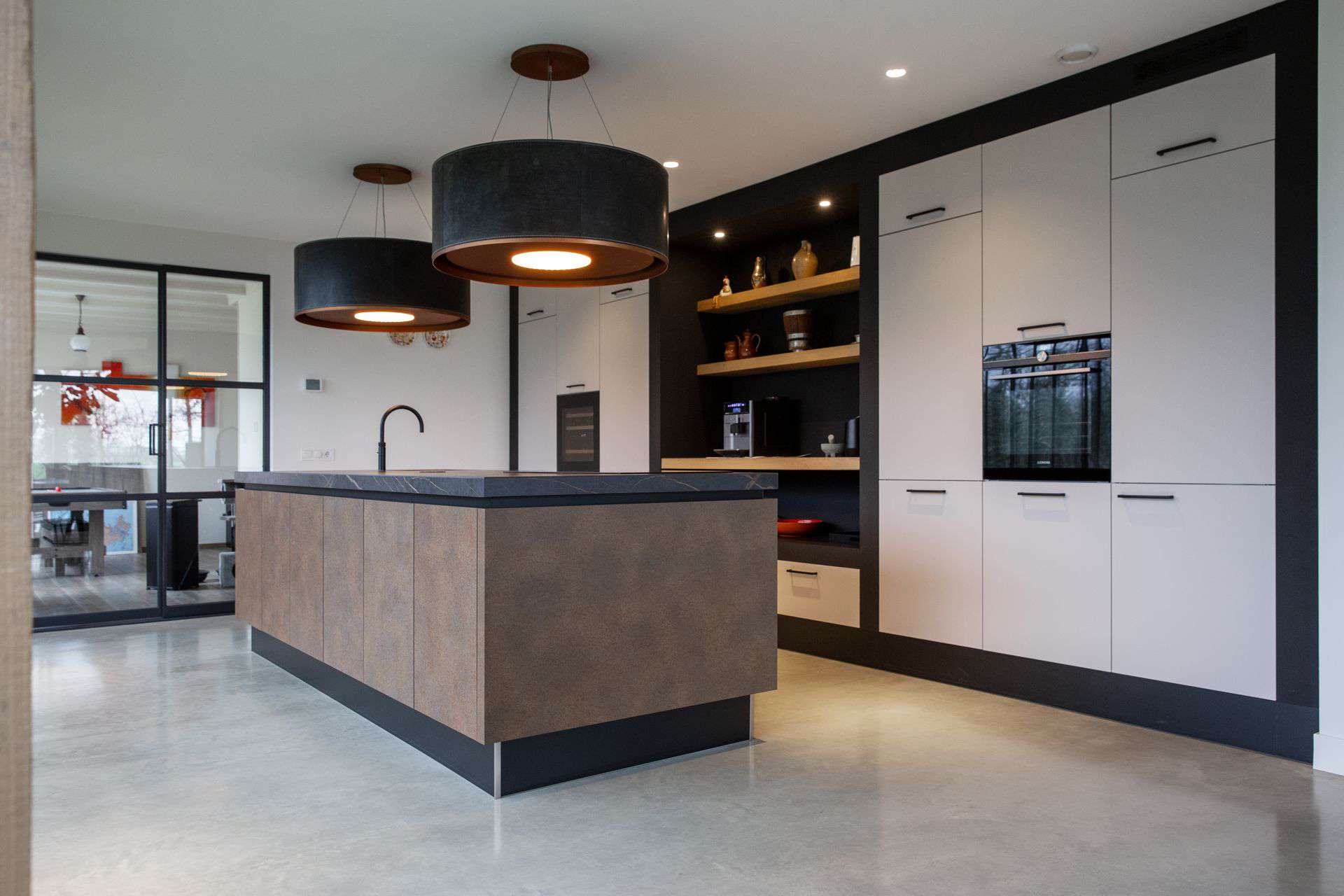 Witte keuken met een bruin kookeiland in Lewedorp. Gecombineerd met een gevlinderde betonvloer en grote zwarte lampen.