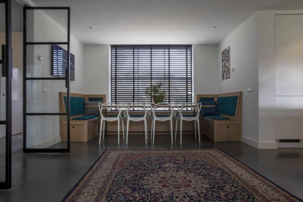 Woning in Beneden-Leeuwen met een gevlinderde betonvloer van Willem Designvloeren in de eetkamer. Dit is gecombineerd met een houten tafel, houten bank en witte stoeltjes. Ook ligt er een gekleurd vloerkleed op de gevlinderde betonvloer.