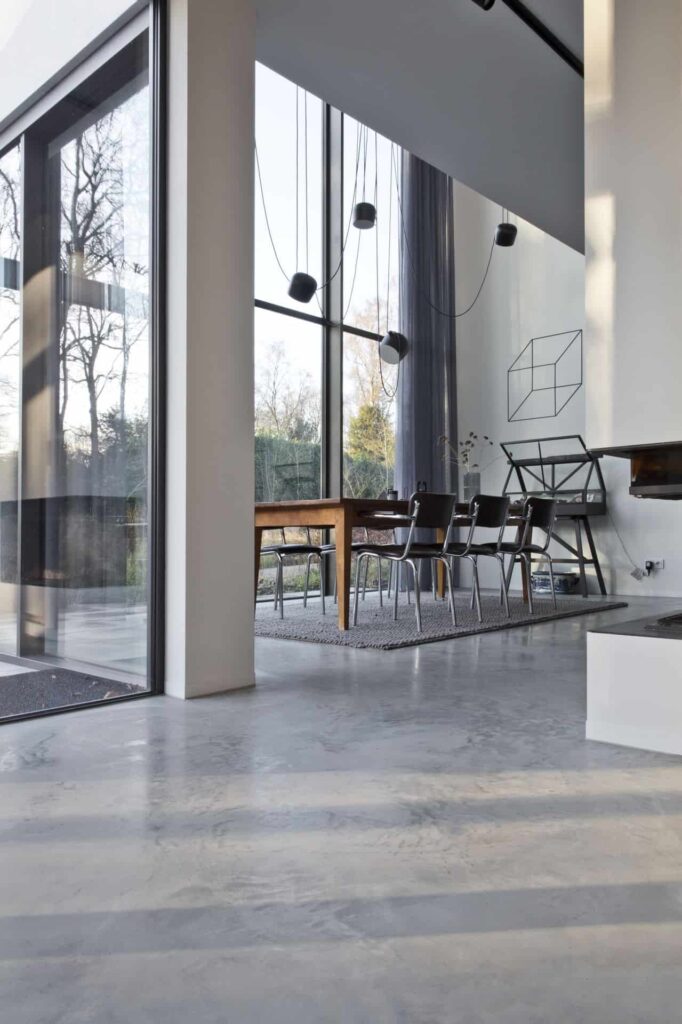 Gevlinderde betonvloer in Huizen met grote raampartijen in de eetkamer. Hier ligt een grijs vloerkleed met daarop een houten tafel en zwarte stoelen.
