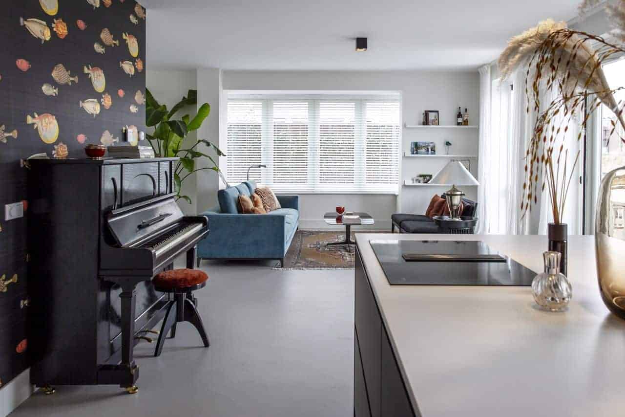 Beton ciré vloer van Willem Designvloeren in de keuken en woonkamer in Kesteren. De vloer is gecombineerd met een blauwe bank en er staat een piano op de vloer.