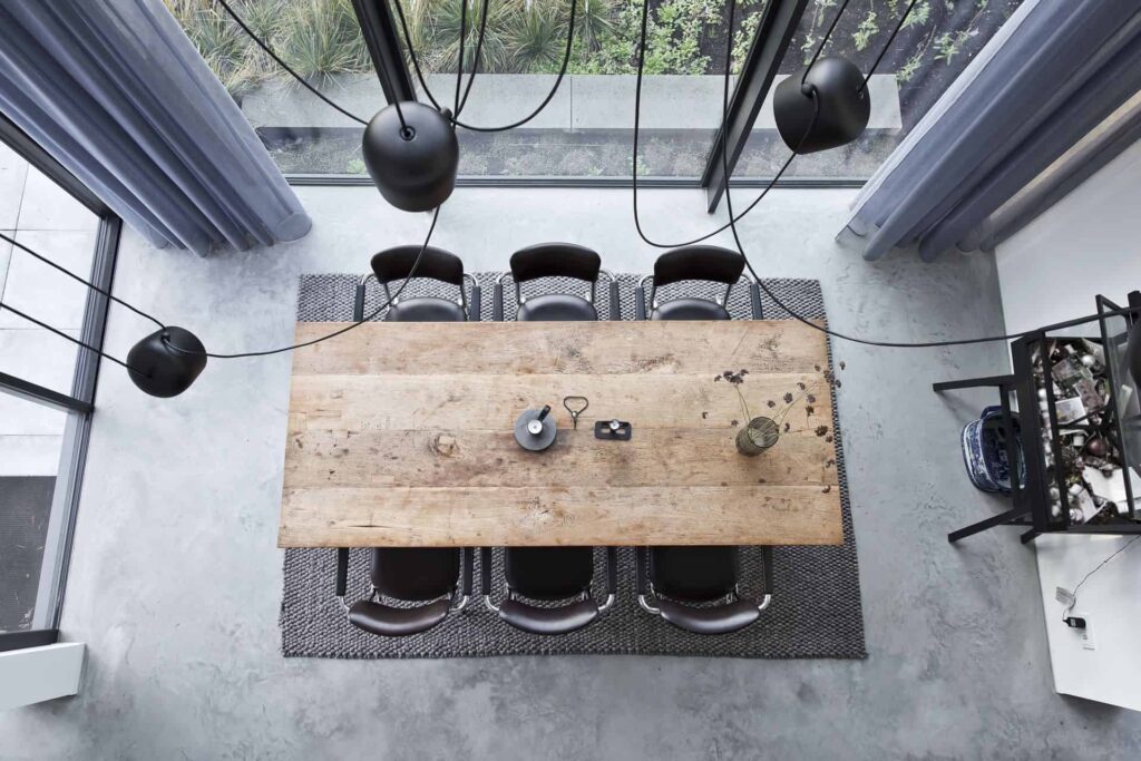 Afbeelding van bovenaf van de eetkamer in Huizen. Hier ligt een gevlinderde betonvloer met daarop een grijs vloerkleed, houten tafel en zwarte stoelen.