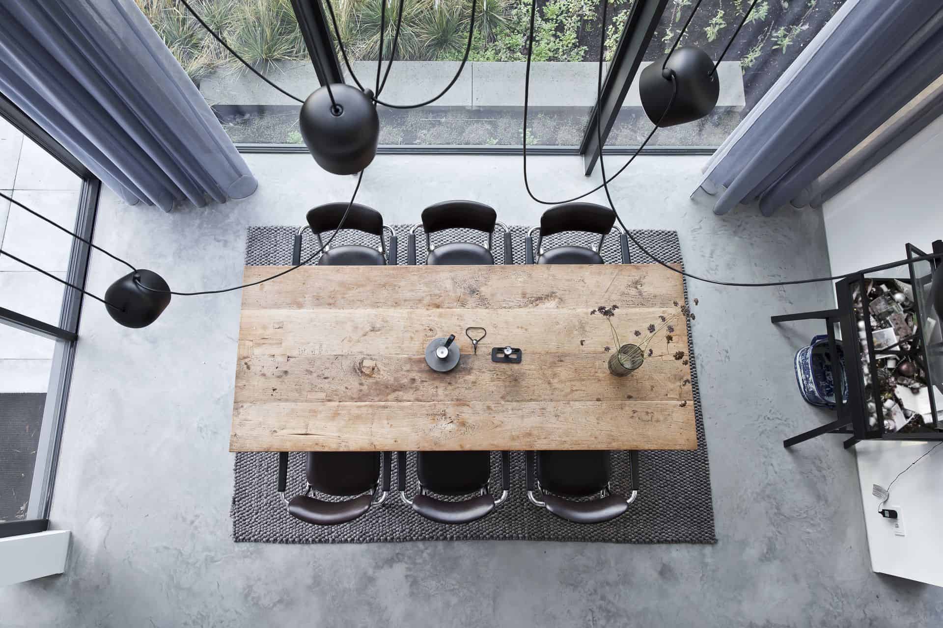 Afbeelding van bovenaf van de eetkamer in Huizen. Hier ligt een gevlinderde betonvloer met daarop een grijs vloerkleed, houten tafel en zwarte stoelen.