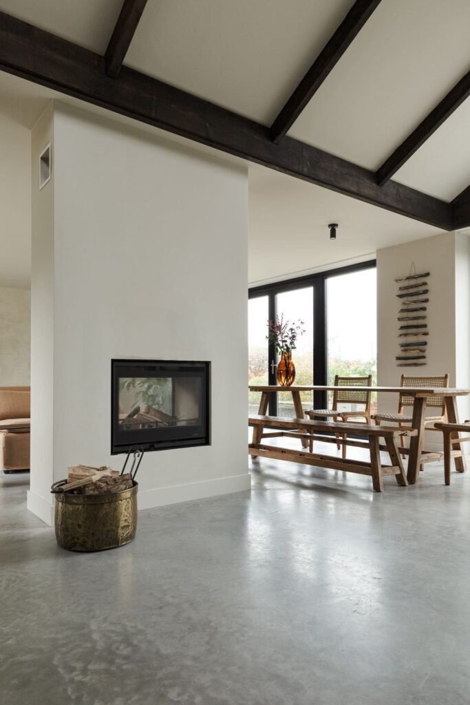 Doorkijk haard in Rijnsburg. Deze staat op een gevlinderde betonvloer. Hiernaast staat een houten eettafel met stoelen en een bankje.