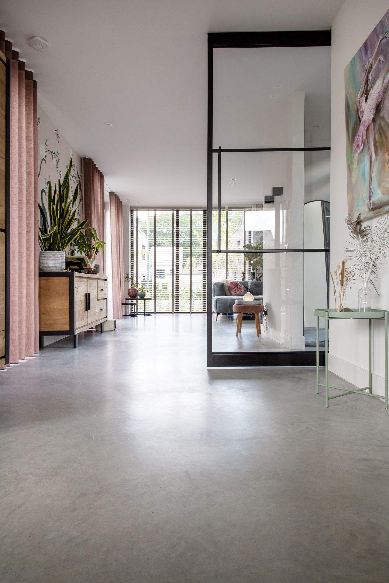 Doorkijkje naar de woonkamer in deze woning in Ede. Hierin is een stalen deur geplaatst, hangen roze gordijnen en is er gekozen voor een gevlinderde betonvloer