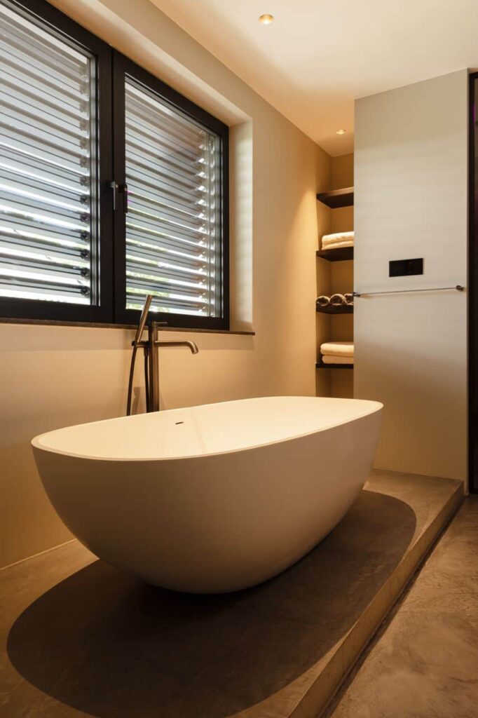 Een ruim opgezette badkamer met een wit vrijstaand bad, zwarte jaloezieën bij de ramen en een prachtige opbergruimte.