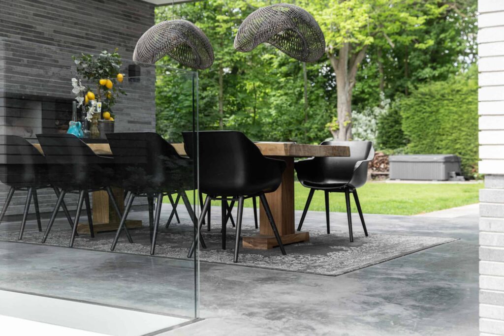 Gevlinderd betonnen terras in Heemstede met hierop een grijs vloerkleed, een houten tafel en 8 zwarte stoelen.