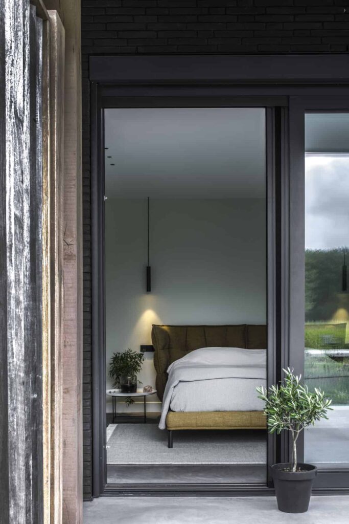 Gevlinderde betonvloer en een gevlinderd betonnen terras in Wijhe dat doorloopt in de slaapkamer. In de slaapkamer ligt een grijs vloerkleed met daarop een groen bed.