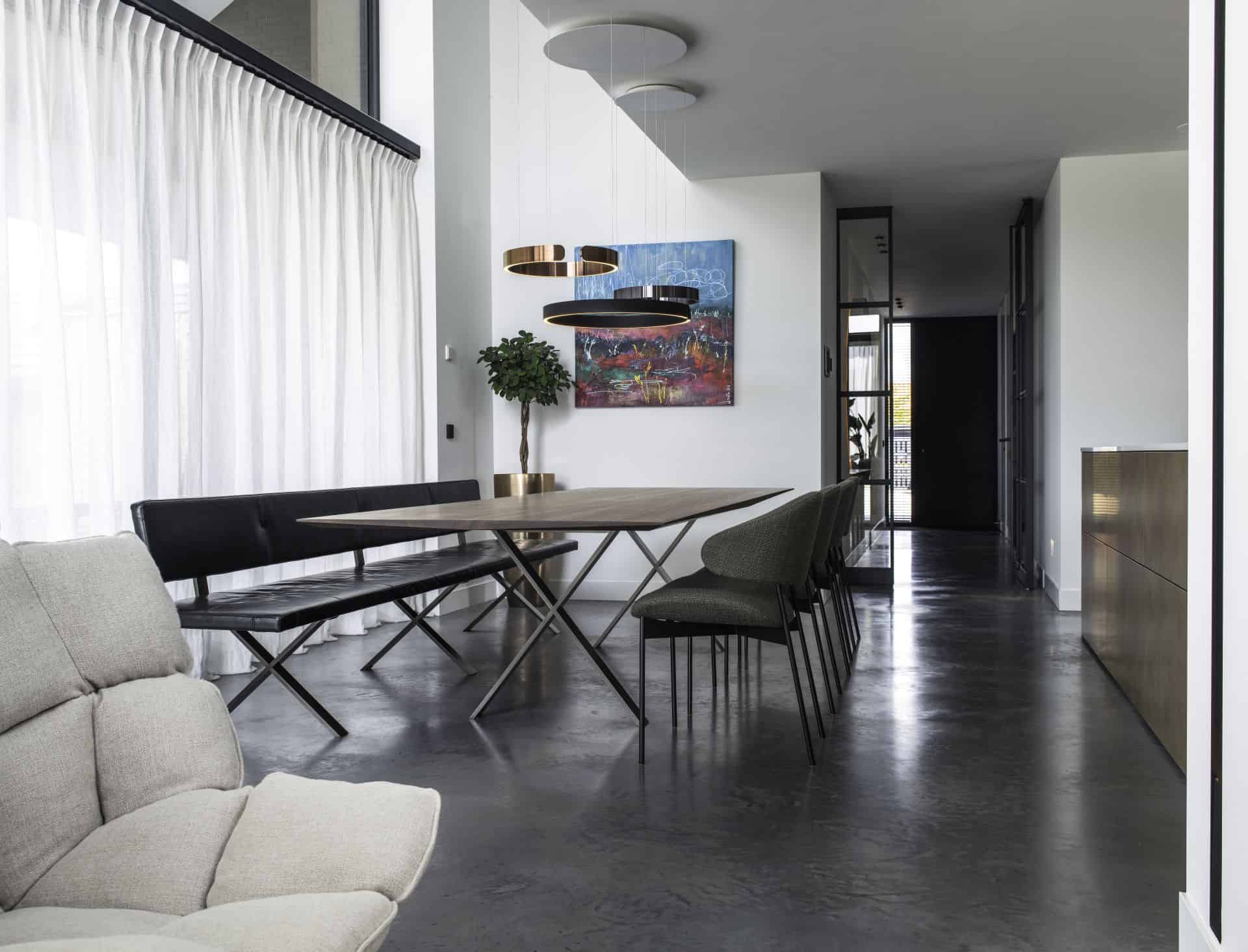 Gevlinderde betonvloer van Willem Designvloeren in Wijhe met daarop een grijze stoel, en een zwarte eettafel met groene stoelen en een zwarte bank.