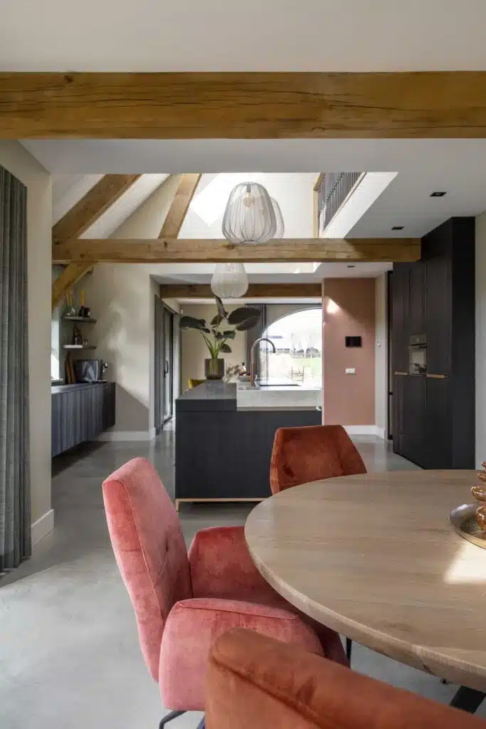 Nieuw gebouwde boerderij in Horst met een gevlinderde betonvloer. In combinatie met een zwarte keuken houten balken aan het plafond een houten tafel met daarom roze stoelen