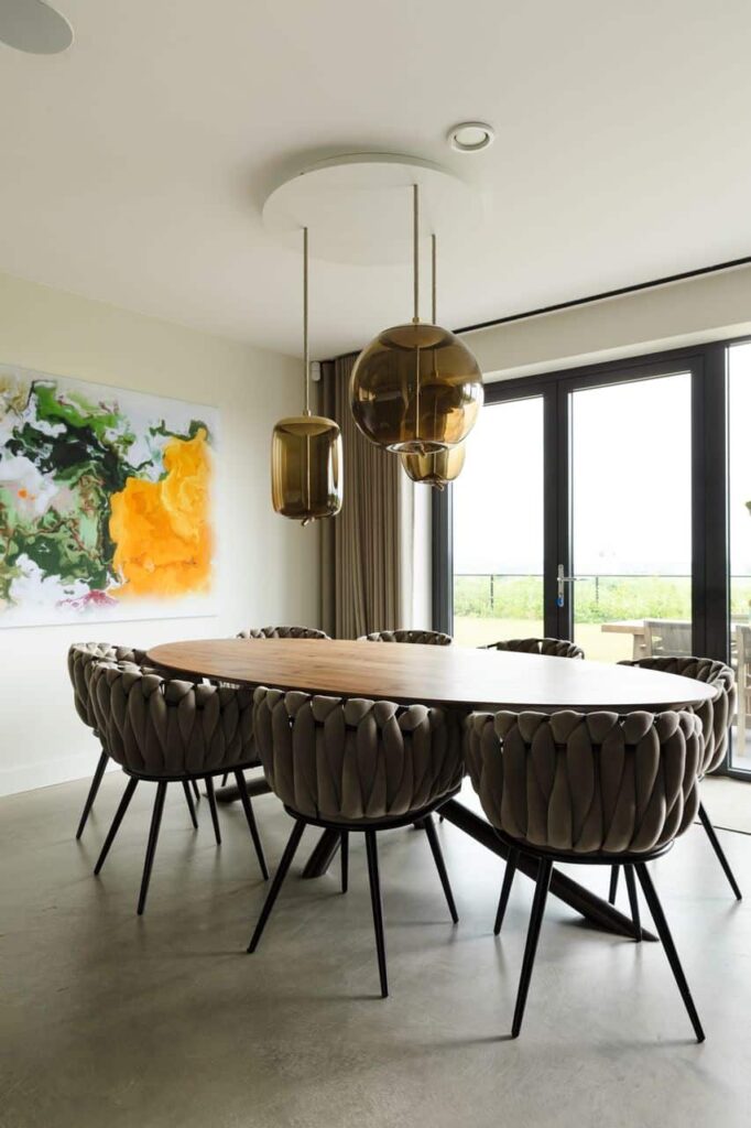 Ovale eettafel met 8 gevlochten kuipstoelen en kleurrijke kunst tegen een witte muur met op de vloer een Italiaanse betonlook vloer.