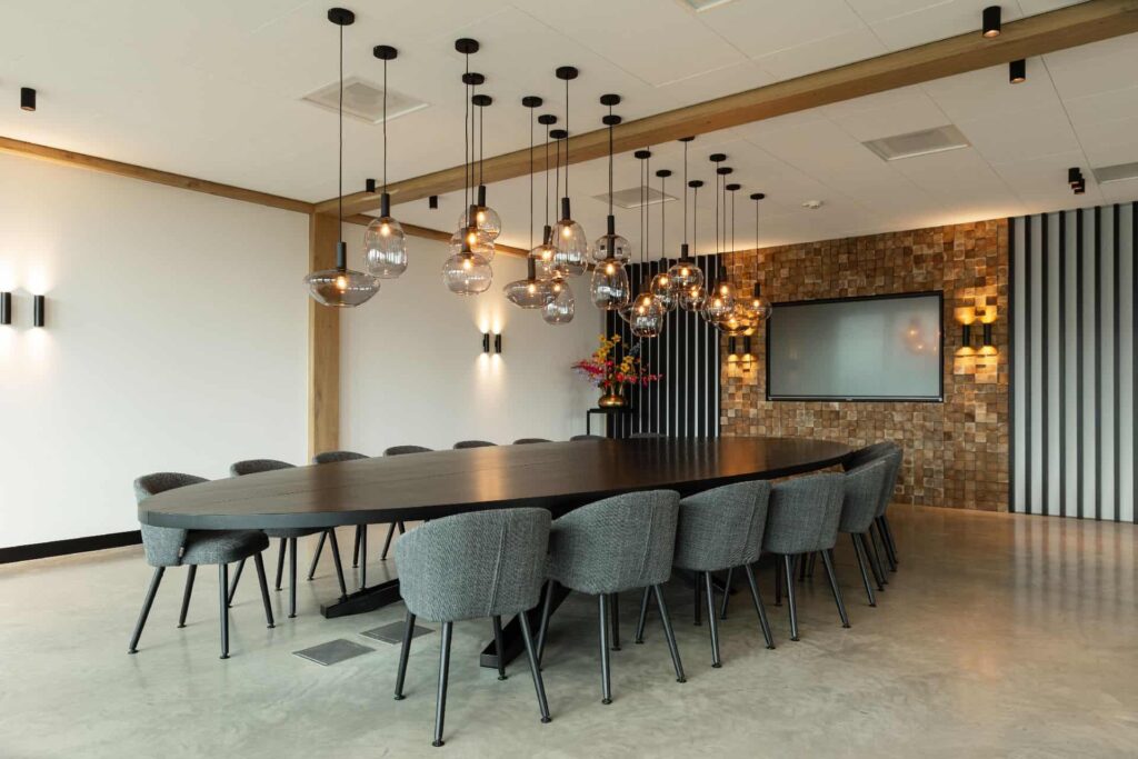 Vergaderruimte in Hattemerbroek met een gevlinderde betonvloer. Hierop staat een grote zwarte tafel met daaraan 16 grijze stoelen. Aan het plafond hangen zwarte lampjes.
