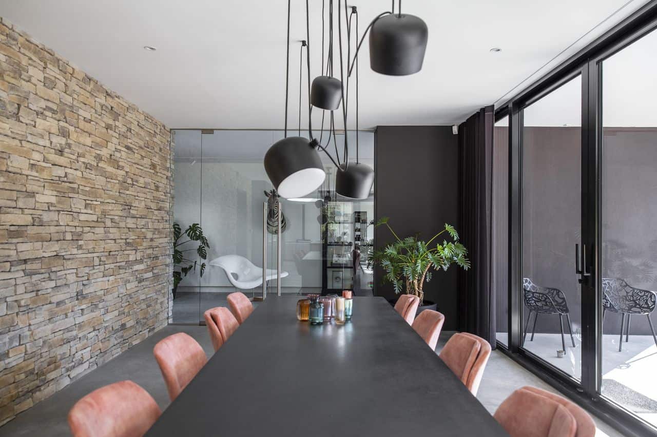 Woning in Nijmegen met een gevlinderde betonvloer van Willem Designvloeren gecombineerd met een stenen muur en een zwart eettafel met hieraan roze stoelen.