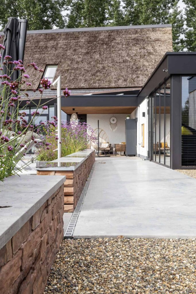 Woning in Nuenen met een gevlinderd betonnen terras. Deze is op afschot gemaakt naar de goot. Hierlangs ligt grind en twee muurtjes met hierachter paarse bloemen.