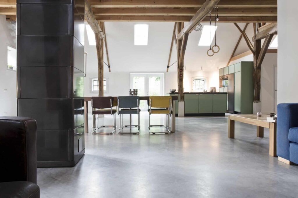 Woon boerderij in Rijswijk met een gevlinderde betonvloer van Willem Designvloeren. Hierop staat een groene keuken en een houten tafel met daaraan verschillende kleuren stoelen.