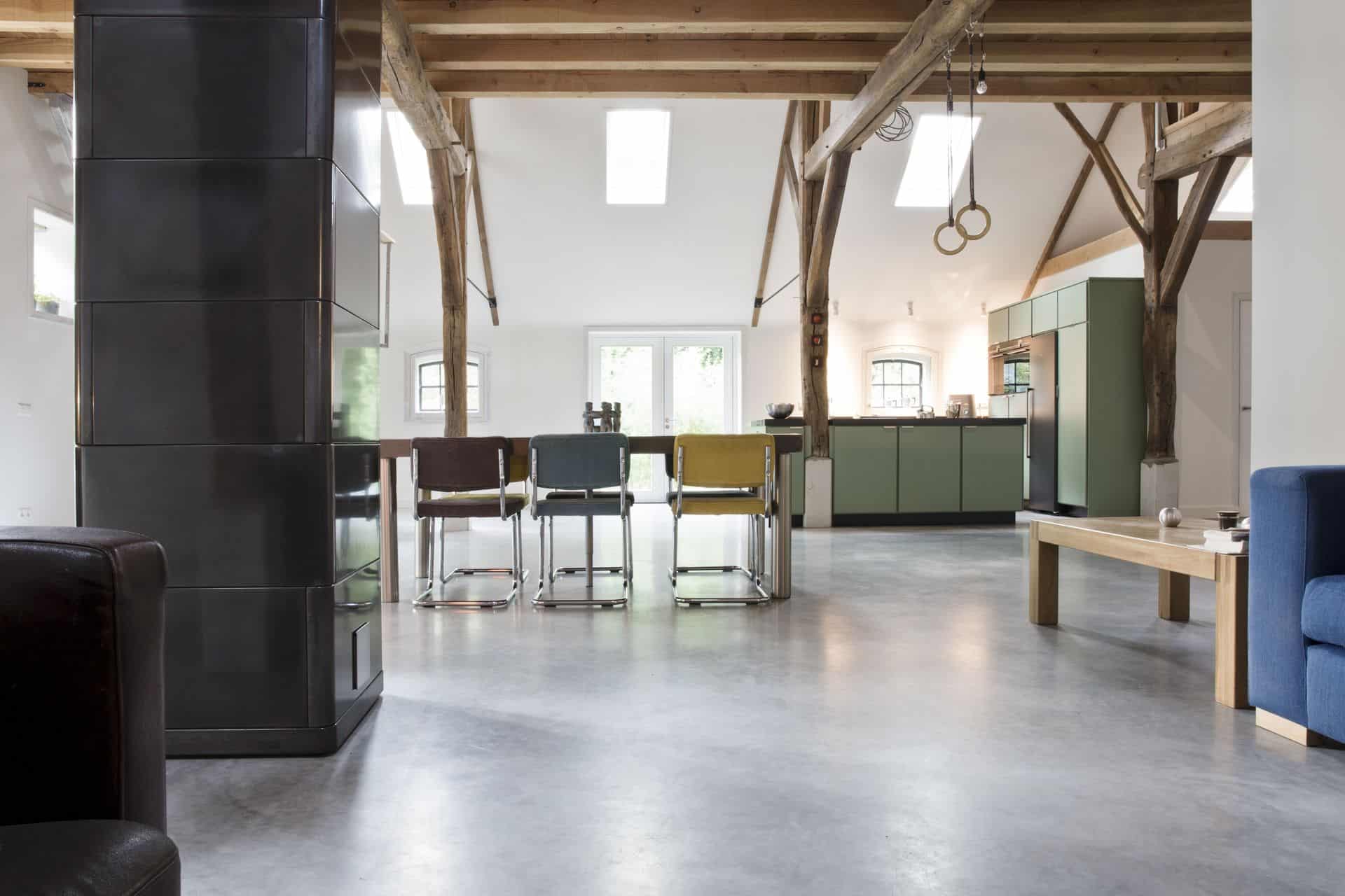 Woon boerderij in Rijswijk met een gevlinderde betonvloer van Willem Designvloeren. Hierop staat een groene keuken en een houten tafel met daaraan verschillende kleuren stoelen.