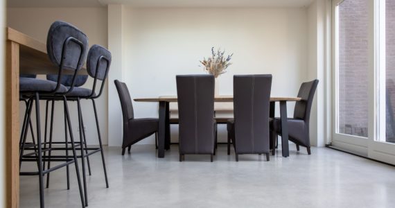 Gevlinderde woonbeton vloer in Sprang-Capelle. Hierop staat een houten eettafel met zwart stalen poten en daaraan 4 zwarte stoelen. Ook zie je een gedeelte van de houten keuken met daaraan blauwe barkrukken.