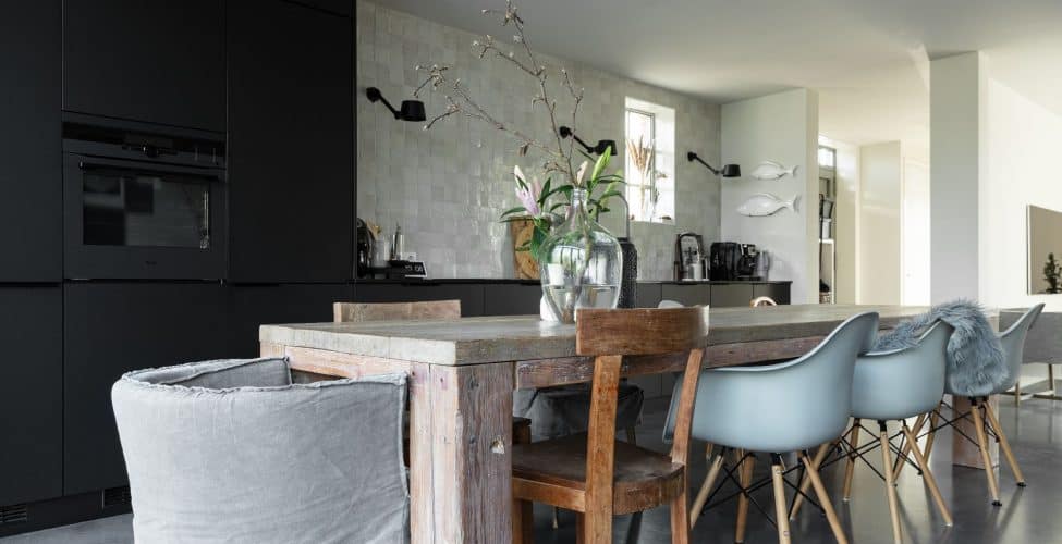 gevlinderde betonvloer gecombineerd met een zwarte keuken, een robuuste eettafel met verschillende stoelen en kleine vierkante tegels als spatwand in de keuken van een vrijstaande Kempische woning.
