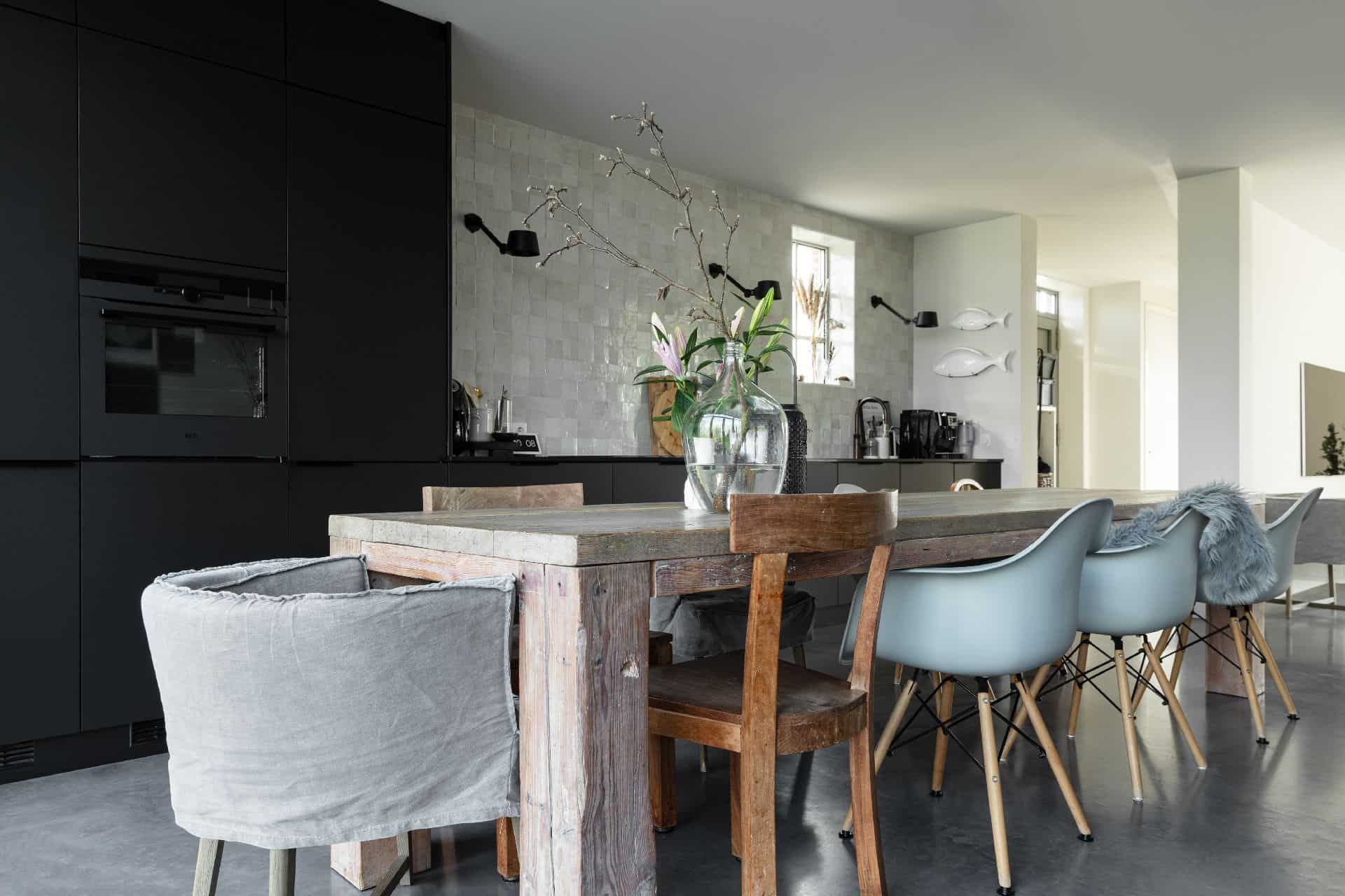 gevlinderde betonvloer gecombineerd met een zwarte keuken, een robuuste eettafel met verschillende stoelen en kleine vierkante tegels als spatwand in de keuken van een vrijstaande Kempische woning.