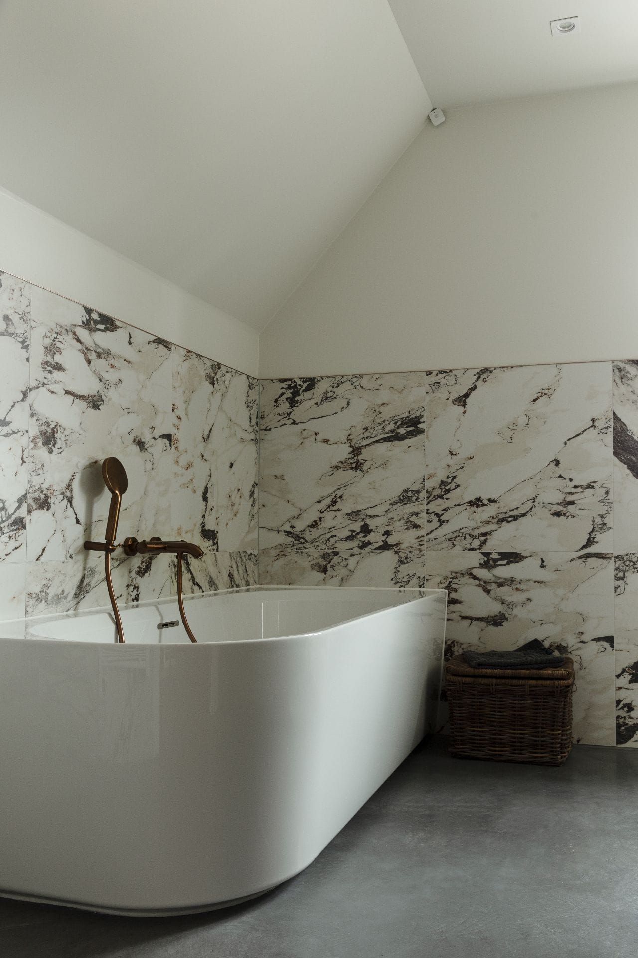 Badkamer met een gevlinderde betonvloer in Brandwijk. Op de vloer staat een wit bad en aan de wand zitten marmeren tegels.