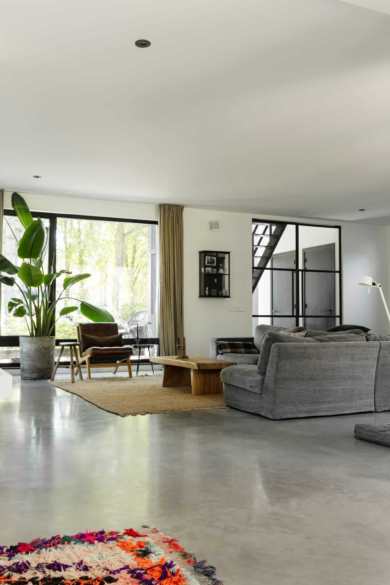 Woonkamer met een grijze bank, houten tafeltje, grote plant, bruine stoel en een gevlinderde betonvloer.