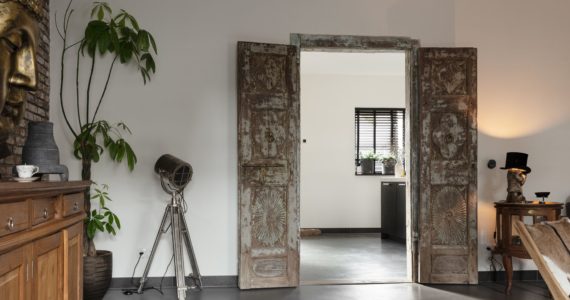 Gevlinderde betonvloer in de woonkamer in Lobith. Deze is gecombineerd met vintage openstaande deuren naar de keuken, een houten kast met hierop een groot buddha beeld en er staat een plant op de vloer.