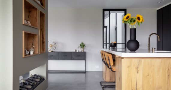 Keuken in Kampen met een gevlinderde betonvloer. De keuken is van hout met hierop een wit blad. Hierachter staat een kast met houten nisjes en daaronder een haard.