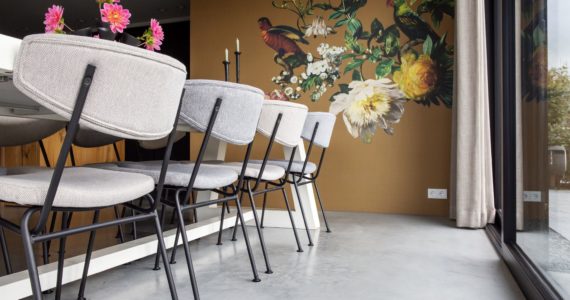 Eetkamer in Kampen met een gevlinderde betonvloer. Hierop staan grijze stoelen met stalen poten. Op de bruine wand zit een muursticker van vogels.