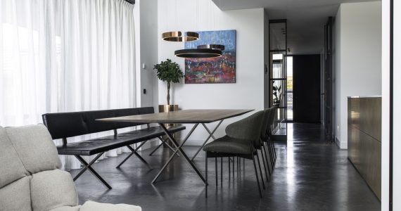 Gevlinderde betonvloer van Willem Designvloeren in Wijhe met daarop een grijze stoel, en een zwarte eettafel met groene stoelen en een zwarte bank.