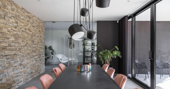 gevlinderde betonvloer in de eethoek, steens muur, roze velvet stoelen, zwarte tafel, hanglampen, glazen pui