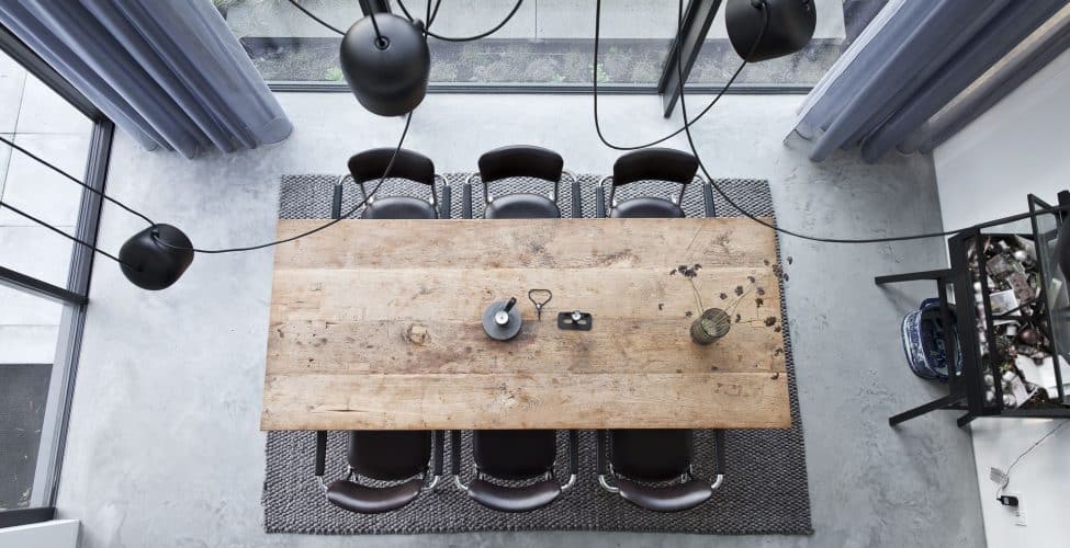 Afbeelding van bovenaf van de eetkamer in Huizen. Hier ligt een gevlinderde betonvloer met daarop een grijs vloerkleed, houten tafel en zwarte stoelen.