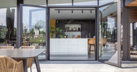 Gevlinderd betonnen terras in Nuenen, gecombineerd met een zwarte tafel. Binnen ligt een gevlinderde betonvloer in de keuken. Hierop staat een wit kookeiland en een zwarte keuken